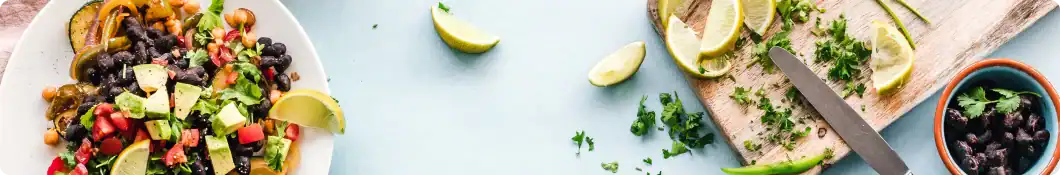 Colorful salad with avocado and black beans next to lemon wedges, chopped herbs, knife, and powder mixer machine.